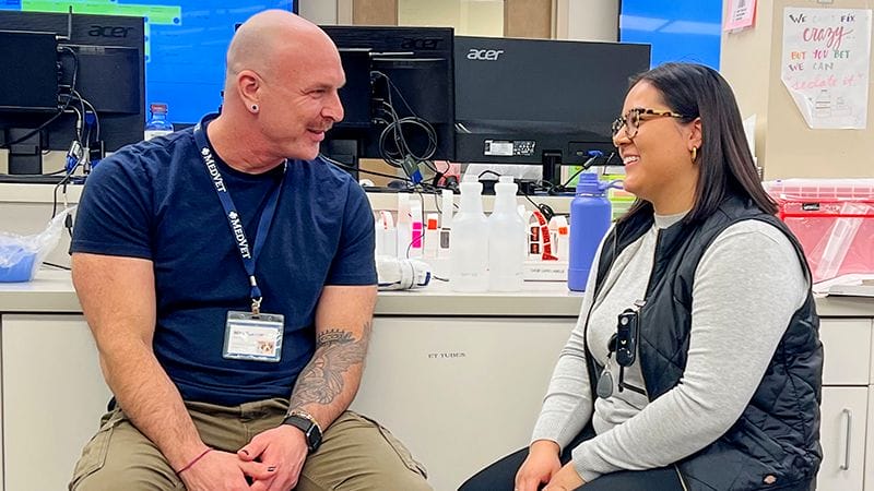 MedVet veterinary social worker in blue shirt smiles as he talks with MedVet team member wearing a grey shirt and black vest